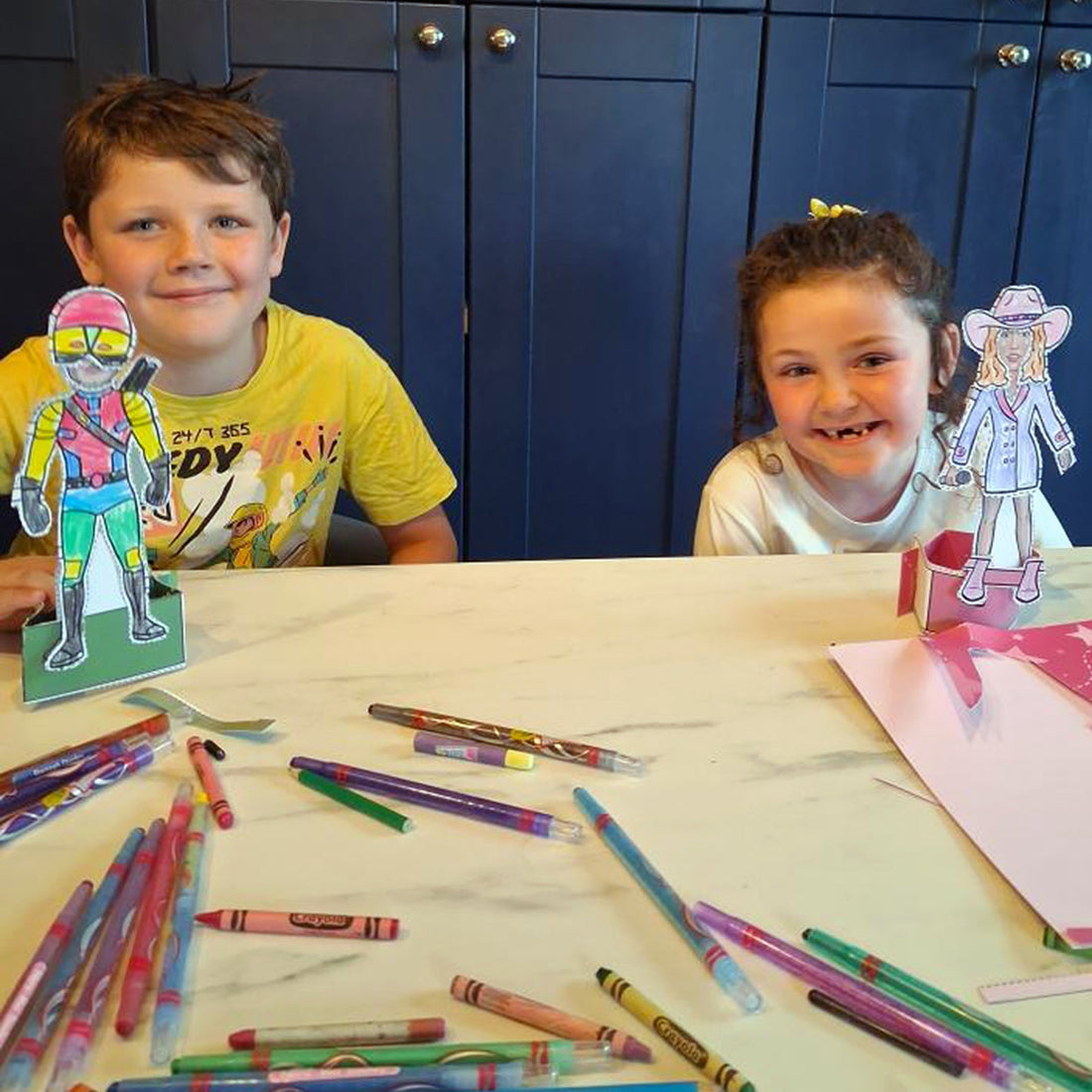 A happy boy and girl sitting at a kitchen table playing with their paper doll books with colouring pencils, markers and crayons on the table.