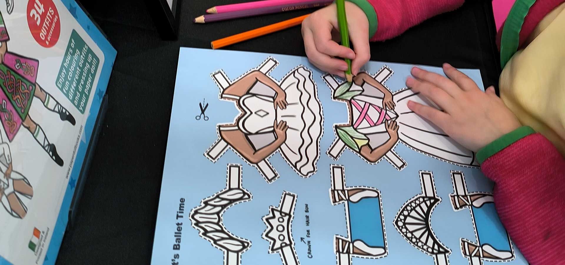 Child coloring in cut-out figures on a blue sheet with colouring pencils in a Paper Doll Book designed and printed in Ireland.