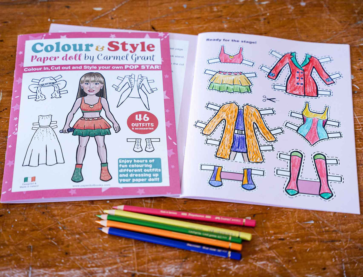 A children's paper doll coloring book by Carmel Grant shown open on a wooden table, with colored pencils placed in front. The left page shows the cover with illustrated paper doll figures and text, and the right page shows colored paper doll outfit templates.