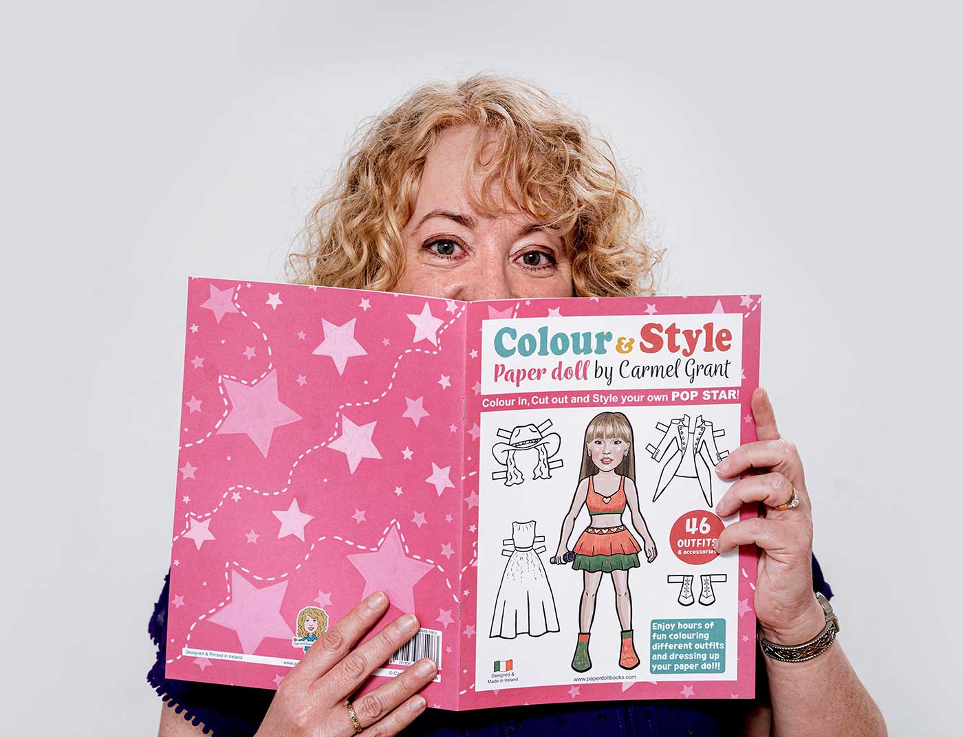 A woman with curly blonde hair holds a pink paper doll book titled 'Colour & Style Paper doll by Carmel Grant' in front of her face against a plain light gray background.