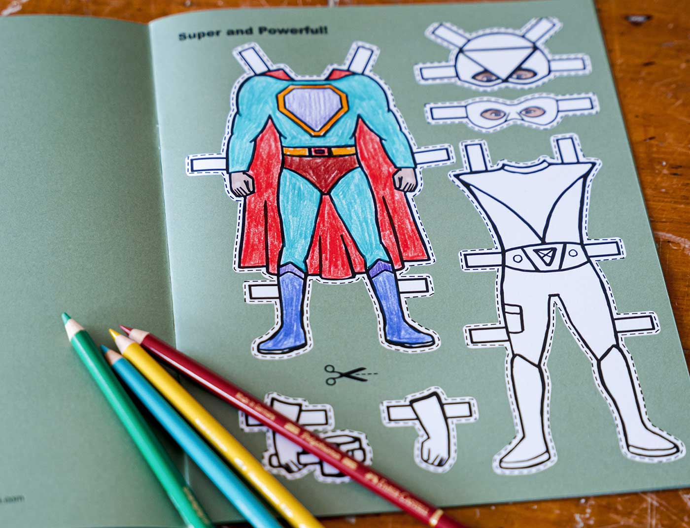 A child's colored superhero paper doll cut out book on a light green sheet, showing a colored superhero figure and an outline of an uncolored body. Three colored pencils are placed on the left side over a wooden table.
