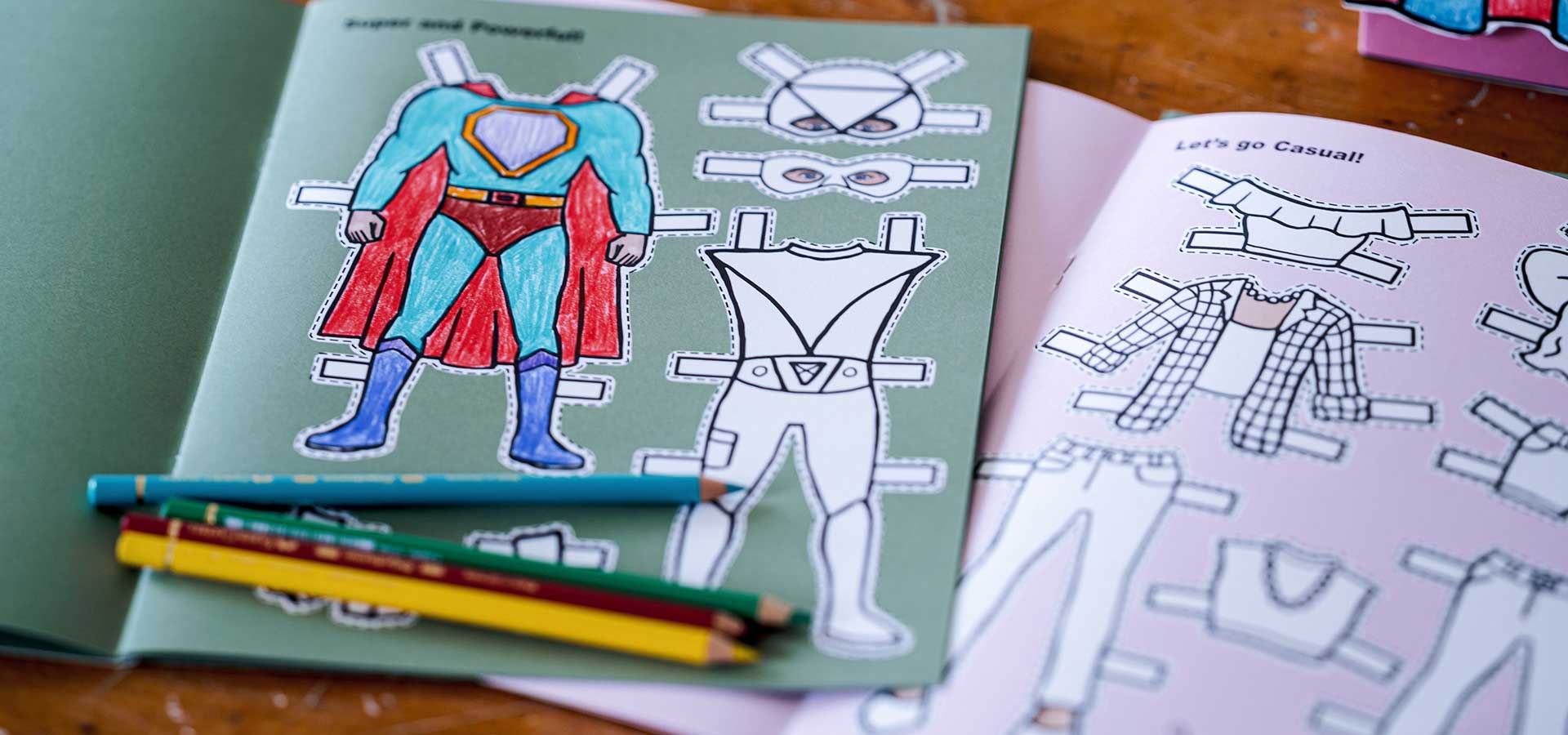 Open pages from two paper Doll cut out books Superhero paper dolls with coloring book and pencils on a wooden surface